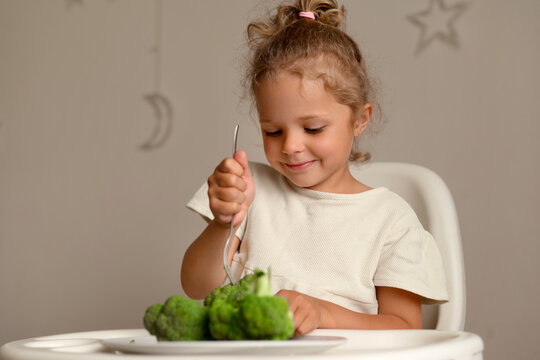 A young child sits at a table in a home and happily uses a fork to eat green vegetables. The setting shows simple decorations and a cozy atmosphere