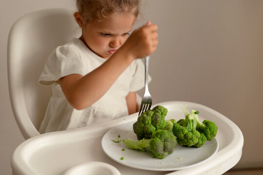 A young child is in a high chair looking frustrated while poking broccoli with a fork. The child appears to dislike the vegetable during lunch at home