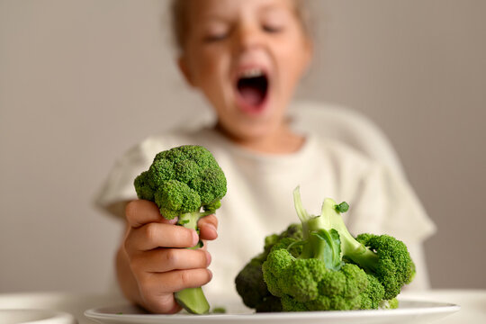 A child is sitting at a table with broccoli. The child yawns while holding a piece of broccoli in one hand. Another piece of broccoli is on the table