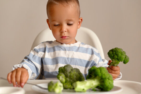 A young child sits at a table with broccoli in front of them. They hold a piece of broccoli while examining the food on their plate
