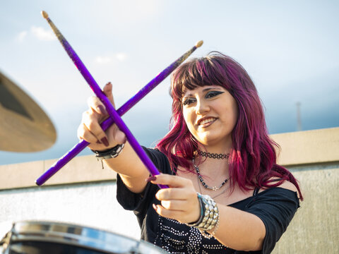 A young female drummer with vibrant purple hair holds drumsticks high in the air while seated behind a drum kit outdoors, expressing confidence, energy, and a passion for music.