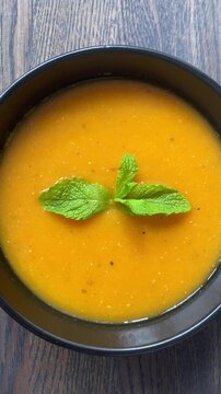 Vertical video. Camera movement. Top view of a black bowl with vegetarian cream soup of red lentils and carrots decorated with mint leaf on a wooden table. Natural ingredients, light food