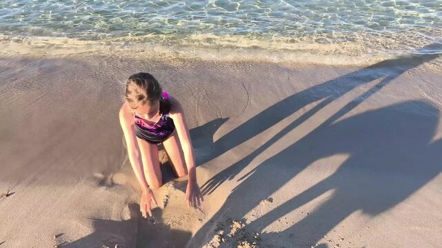 Young girl playing in the sand at the seashore as gentle waves wash onto the beach. Summer vacation, childhood fun, family travel, and outdoor recreation by the sea.