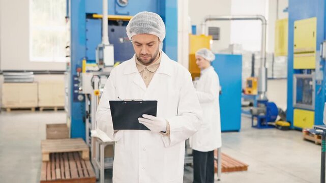 Handsome Caucasian male worker wearing lab coat and protective cap holding clipboard and pen. Looking at large industrial machinery and filling out production data. Colleges working at the background.