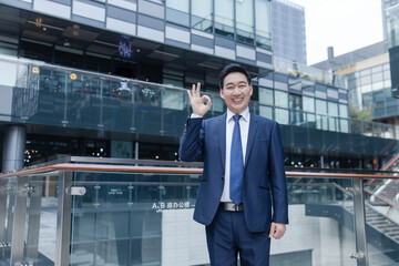 Fototapeta premium Smiling businessman showing OK hand gesture outside modern office building