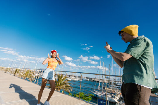 Young man filming happy woman enjoying music at harbor