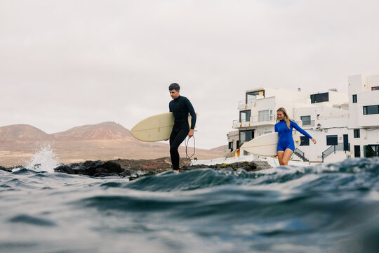 A man and a blonde woman carry surfboards along a rocky shore.