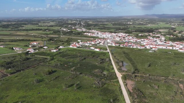 Aerial drone shot pushing in over the picturesque village of Rogil, Portugal. Surrounded by lush green fields and distant hills, showcasing the serene landscape of the Fisherman's Trail.