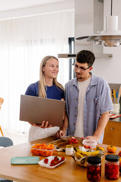 A woman with a laptop smiles at a man standing by a kitchen island.