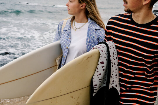 A couple walks along the coast, each carrying a surfboard underarm.