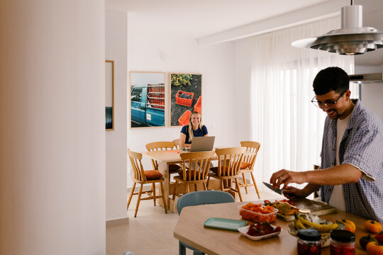 A man cooks at an island while a blonde woman uses a laptop behind.