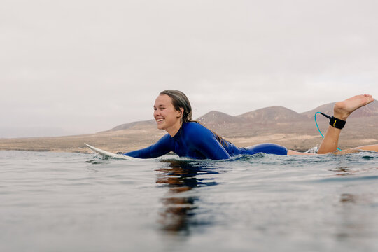 A smiling blonde woman in a blue wetsuit paddles on her surfboard.
