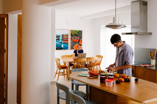 A man prepares food while a blonde woman works on her laptop behind.