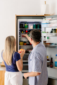 A man reaches into a fridge while a woman holds a fruit container.