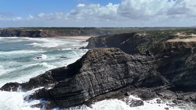 An aerial drone shot pans left over the stunning, wild coastline of Portugal, revealing dramatic cliffs, a sandy beach, and crashing ocean waves.