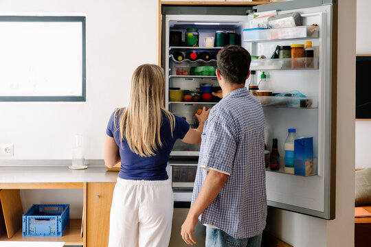 A couple looks inside an open refrigerator in a bright kitchen.