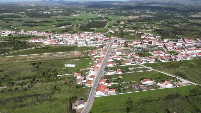 Aerial pan left over Rogil, a charming village nestled amidst lush green fields and rolling hills in rural Portugal, showcasing its houses and peaceful surroundings.