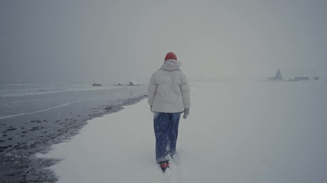 Person walking along snowy shoreline wearing red hat