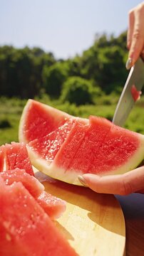 Vertical video. Woman Transferring Fresh Watermelon Cubes From the Rind With a Kitchen Knife on a Wooden Board Outdoors, Close Up, Healthy Summer Snack