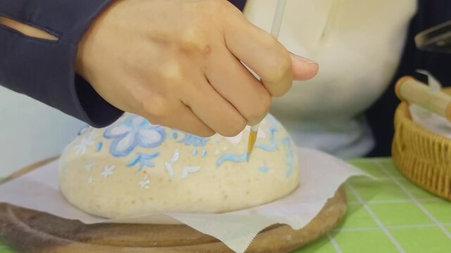 Baker's hands brushing flour onto shaped sourdough bread dough in a Dutch oven, close-up of artisan home baking preparation before scoring, rustic dark kitchen