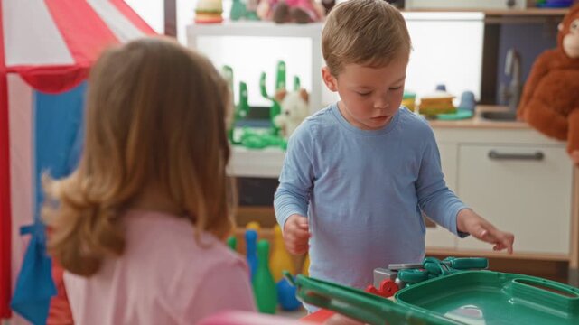 Boy holding a toy screwdriver, turning it with his hand in a building playroom among colorful toys while a girl watches nearby; concentration.