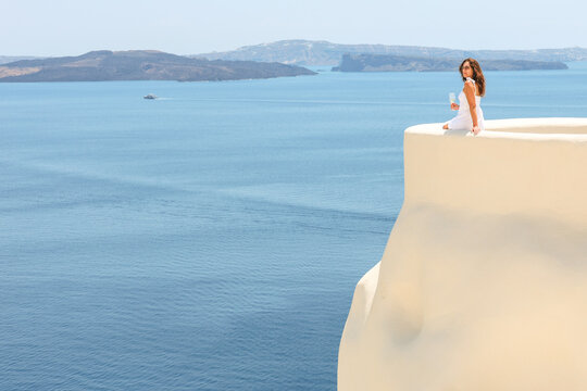 Golden-hour sunshine serenity above the Santorini caldera as a beautiful woman in a white dress enjoys a chilled glass of white wine, soaking in the sweeping Aegean view.