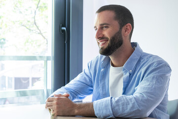 Fototapeta premium Smiling young man in a blue shirt sitting by a window, relaxed and thoughtful in a bright modern home or office setting