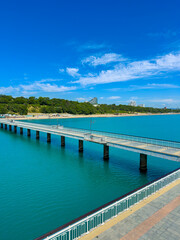 Fototapeta premium Pier extends into blue water under clear sky near green shore with buildings in the distance on a sunny day