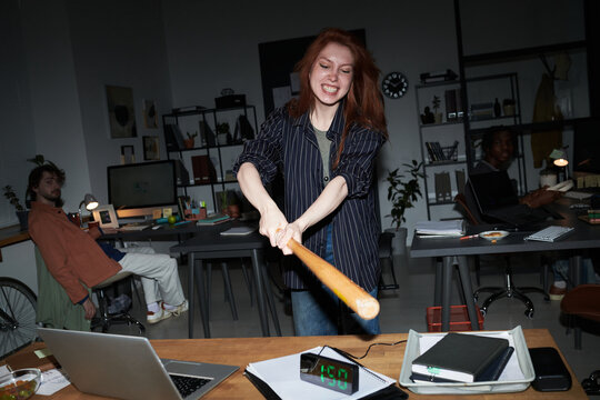 Young adult Caucasian woman smashing digital clock with baseball bat in office, expressing workplace stress and burnout while coworkers watching. For business pressure, anger management, toxic culture