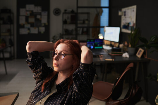 Young adult Caucasian businesswoman leaning back in office chair, pausing during computer work and reflecting on workload. Useful for burnout, remote work, stress management marketing