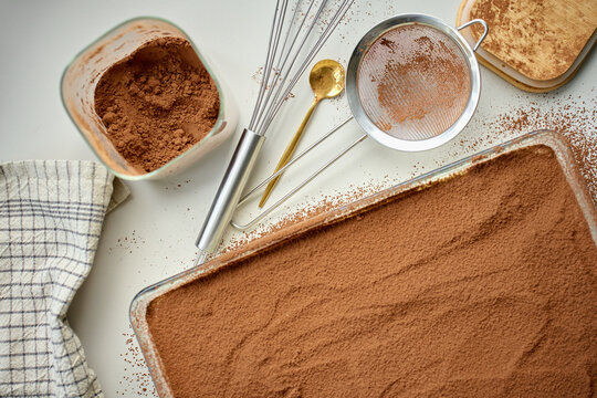 Cocoa covered tiramisu sits in rectangular glass dish beside sieve and cocoa bowl on table