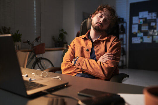 Young adult Caucasian man sleeping in office chair beside laptop, showing overtime fatigue and workplace burnout. Useful for remote work stress, productivity loss, employee wellness marketing