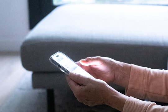 Senior woman using smartphone with both hands while seated on a couch, focusing on screen light and touch interaction, illustrating aging, connection, communication and everyday technology.