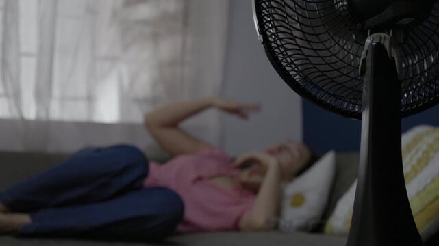 Electric fan cooling a woman resting at home during an extreme summer heat wave. Indoor comfort and climate change concept.