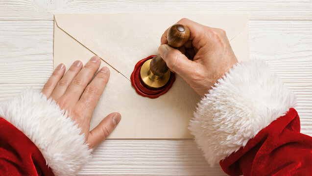 Santa Claus hands pressing red wax seal on Christmas envelope over white wood table. Concept of Santa letter, holiday greeting, festive mail and seasonal ad