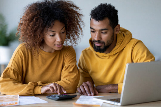 Couple in matching yellow sweaters examines finances together. They focus intently on laptop and calculator. Their calm expressions show thoughtful collaboration