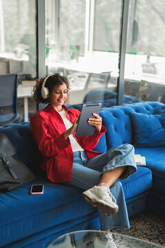Young latin or indian female student studying on digital tablet technology in headphones for online learning. Vertical portrait of woman browsing educational content on device in campus or office.