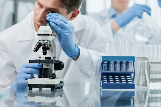 Young scientist or researcher in protective workwear studying new virus in microscope to develop antidote for fighting coronavirus