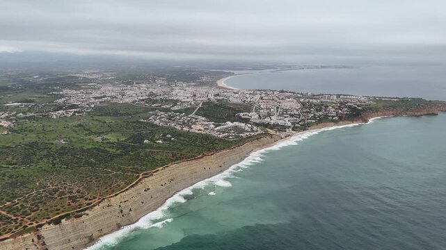 Aerial pan right view of the dramatic coastline, cliffs, and sandy beaches near Lagos, Portugal. The city blends into the lush landscape, part of the Fisherman's Trail on the Rota Vicentina.