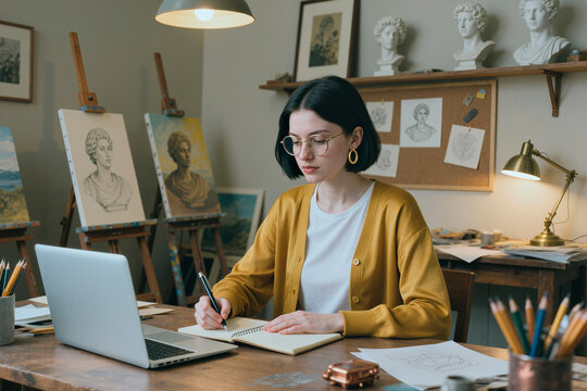Young adult Caucasian woman sketching classical sculpture studies beside laptop in art studio, developing creative portfolio. Ideal for art education, online learning, creative career content