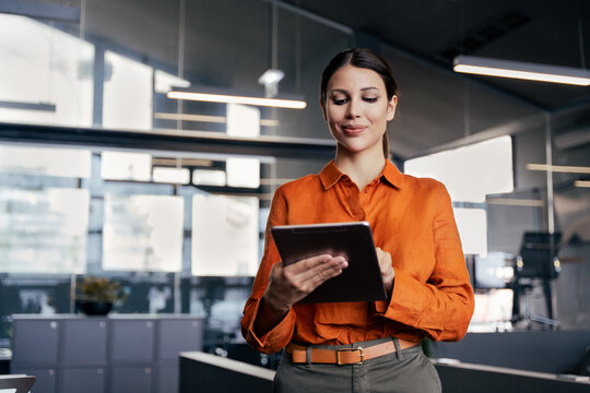 Young latin or indian businesswoman using digital tablet standing in modern office. Confident professional woman checking data, browsing online application in corporate workplace, business technology.