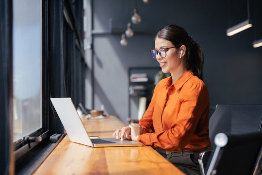 Smiling young latin or indian businesswoman wearing glasses working on laptop computer at wooden desk by window. Professional woman typing, listening music in earphones in modern coworking office.