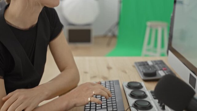 Woman resting chin on hand while pausing from typing at computer keyboard beside microphone and green screen studio backdrop; pensive concentration.