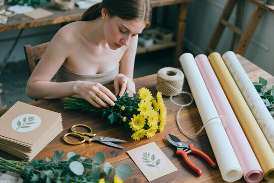 Young adult Caucasian woman arranging flower bouquet and wrapping stems at worktable, creating handmade floral product. Ideal for small business, craft branding, retail marketing