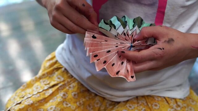 Girl&rsquo;s paint-stained hands unfolding a handmade watercolor fan painted in watermelon colors. The fan opens into a semicircle against the background of a yellow floral skirt and white T-shirt.
