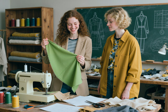 Young adult Caucasian women examining fabric sample in fashion design studio, collaborating on garment development. Ideal for apparel education, tailoring business, creative teamwork