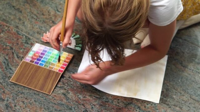 girl sitting on a stone floor, using a wide brush to paint  a sheet with golden-yellow watercolor containing shimmer particles. Next to her lie a colorful watercolor palette and a folded handmade fan 
