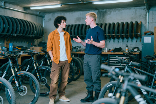 Young adult men discussing bicycle options in repair shop, comparing models and purchase needs. Useful for cycling retail, customer service, maintenance, sales marketing