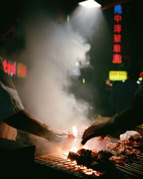Street food vendor grilling fresh meat skewers over hot open charcoal flames during a busy night in a vibrant city