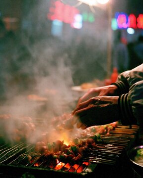 Street food vendor skillfully grilling delicious meat skewers over a hot charcoal fire during a busy colorful night market scene